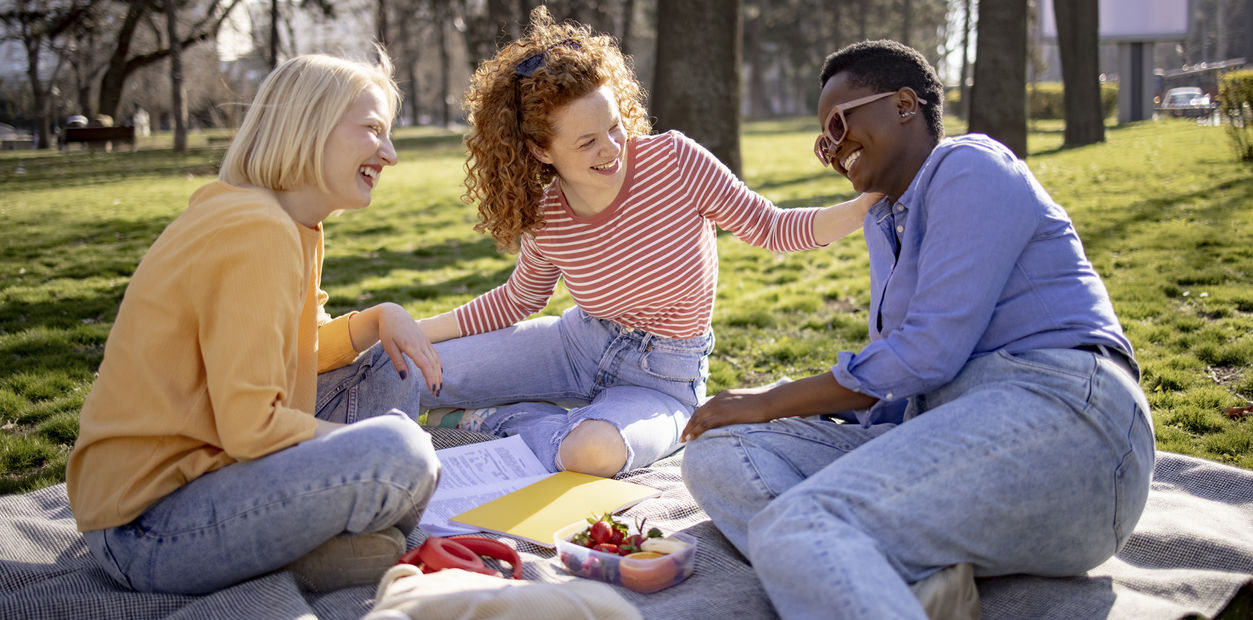 image of three female students having a picnic outdoors. They are smiling at one another and have snacks and books between them.